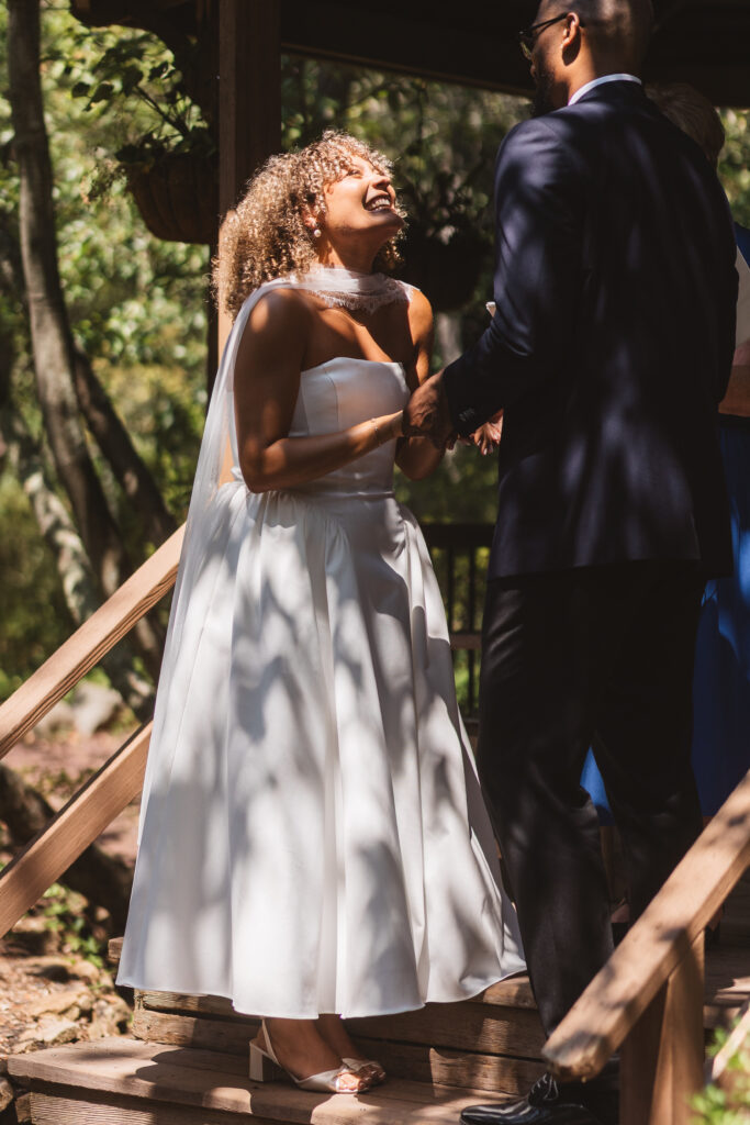 Bride smiles up at her groom as they hold hands and are partially shaded by the trees in Sayen Gardens, photographed by Philadelphia and New Jersey based travel wedding photographer, Madison Neumann Photography.