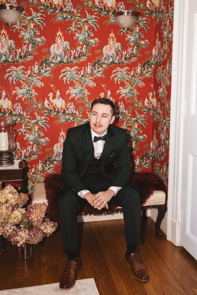 Portrait of groom sitting on a bench in front of a beautiful red decorated wallpaper, photographed by Philadelphia and New Jersey based travel wedding photographer, Madison Neumann Photography.