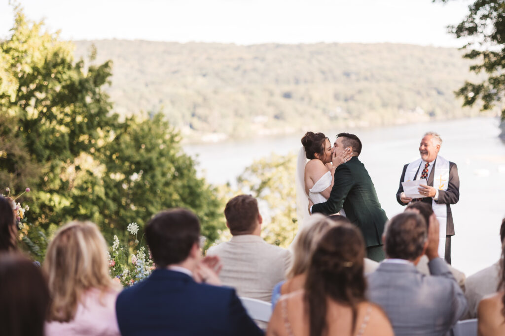 First kiss as husband and wife with happy guests celebrating with them overlooking the water at St. Clements Castle, photographed by Philadelphia and New Jersey based travel wedding photographer, Madison Neumann Photography.