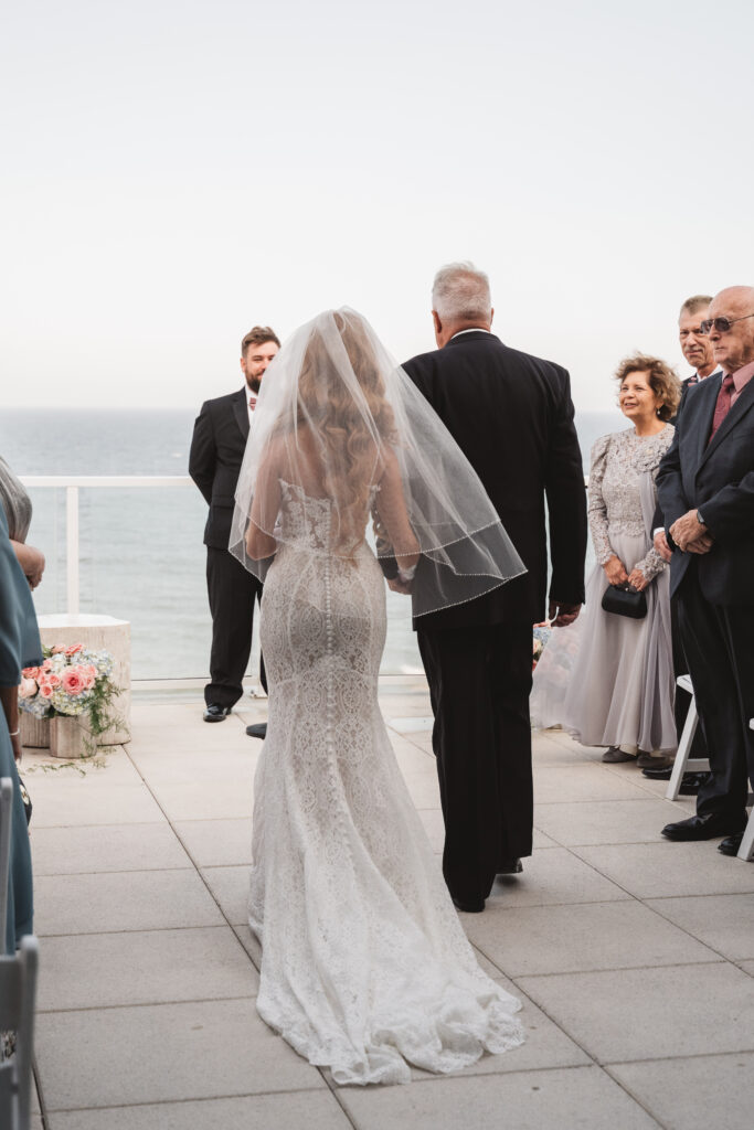 Shot from behind the bride as she walks down the aisle with her father overlooking the ocean at Wave Resort, photographed by Philadelphia and New Jersey based travel wedding photographer, Madison Neumann Photography.