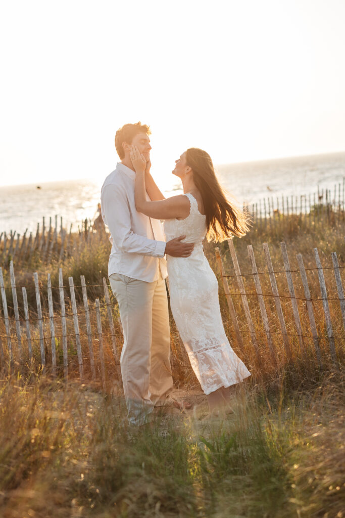 Happy couple is on the beach as the sun goes down, future Mrs. holds her fiancé's face lovingly and smiles at him as the sun peaks behind them, photographed by Philadelphia and New Jersey based travel wedding photographer, Madison Neumann Photography.
