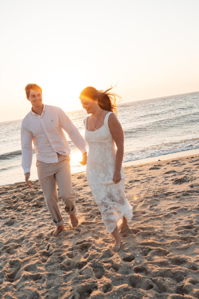 Happy couple running on the beach together as the sun peaks out behind them, casting a beautiful orange glow, photographed by Philadelphia and New Jersey based travel wedding photographer, Madison Neumann Photography.