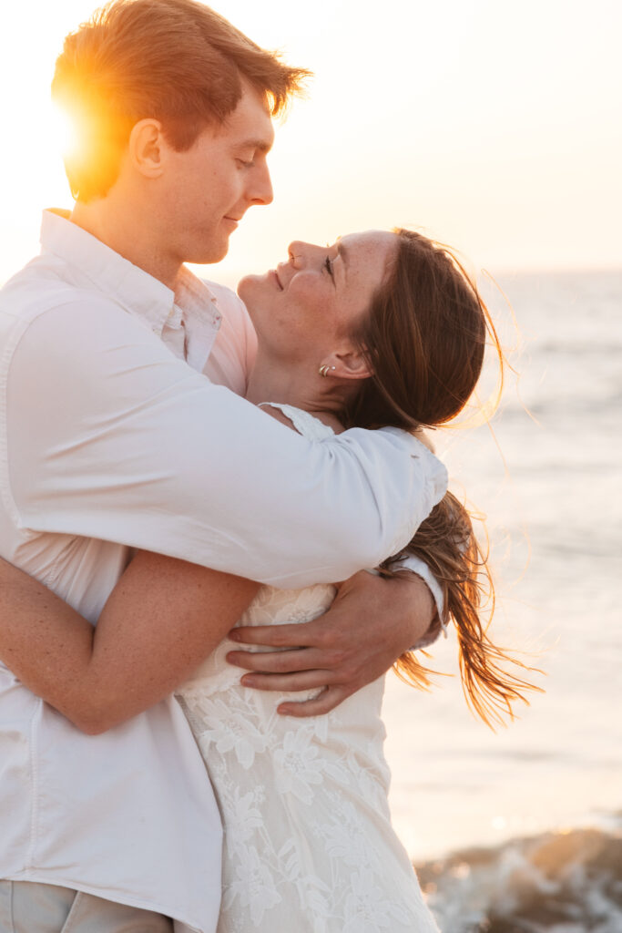 Happy couple shares a heartfelt hug on the beach with the sun peaking behind the grooms head as they smile contently at each other, photographed by Philadelphia and New Jersey based travel wedding photographer, Madison Neumann Photography.