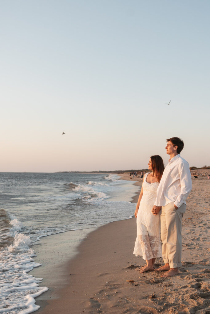 Future Mr. and Mrs. hold hands at the waters edge and watch the sunset together as washes crash and birds fly around them on the beach in Cape May, NJ. Photographed by Philadelphia and New Jersey based travel wedding photographer, Madison Neumann Photography.