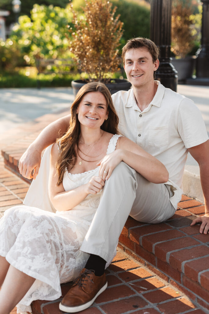 Future bride sits between her fiance's legs on red brick steps in Cape May New Jersey, photographed by Philadelphia and New Jersey based travel wedding photographer, Madison Neumann Photography.