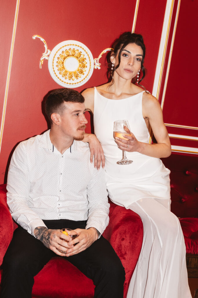 Groom sits in a red velvet chair as his fiancé sits on the arm with her hand on his shoulder while holding their drinks, photographed by Philadelphia and New Jersey based travel wedding photographer, Madison Neumann Photography.