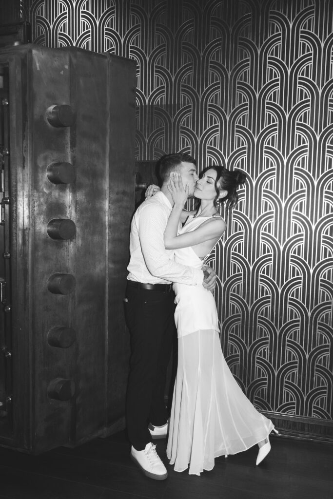 Black and white photo of future groom holding his fiancé's waist and kissing her on the cheek with a 1920s type wallpaper behind them, photographed by Philadelphia and New Jersey based travel wedding photographer, Madison Neumann Photography.