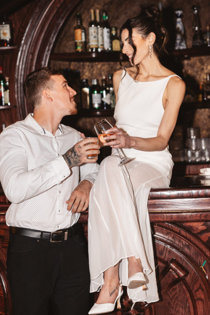 Bride sits on the bar as her fiancé stands next to her as they cheers their drinks in celebration, photographed by Philadelphia and New Jersey based travel wedding photographer, Madison Neumann Photography.
