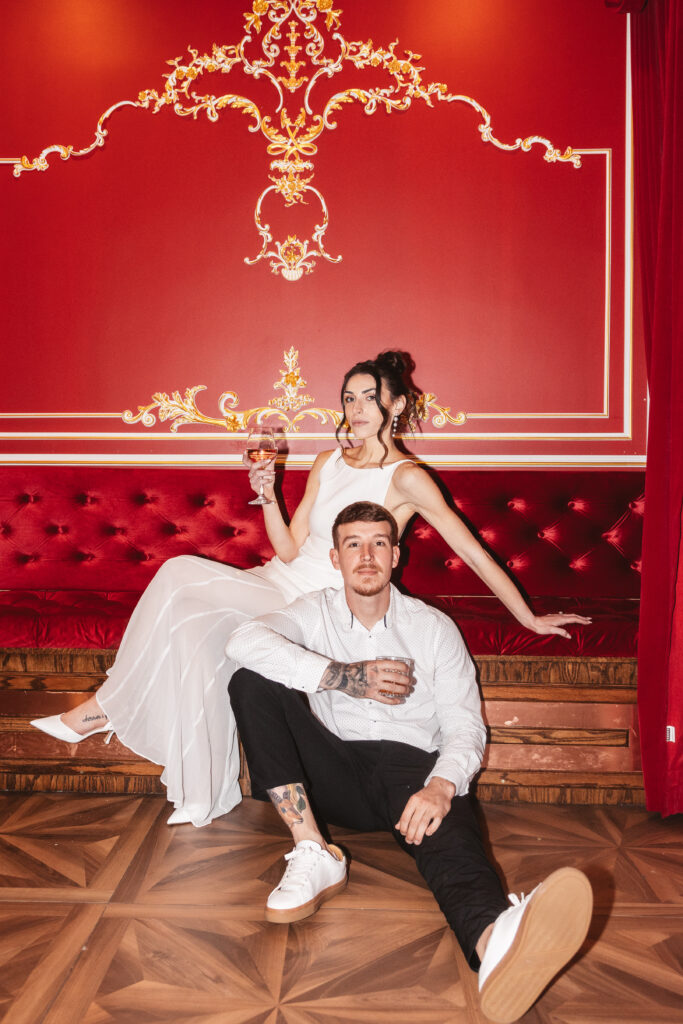 Groom sits on the floor while the bride lays comfortably on a red velvet built-in bench with red wallpaper and gold detailing, photographed by Philadelphia and New Jersey based travel wedding photographer, Madison Neumann Photography.