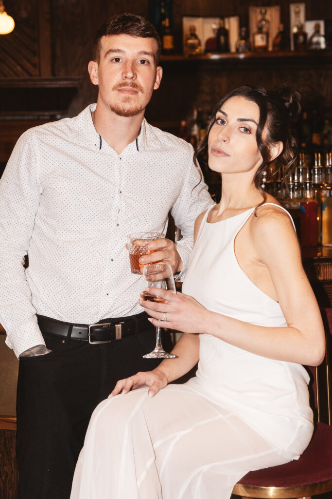 Bride sits on bar stool and her fiancé stands beside her, both with drinks in hand, photographed by Philadelphia and New Jersey based travel wedding photographer, Madison Neumann Photography.