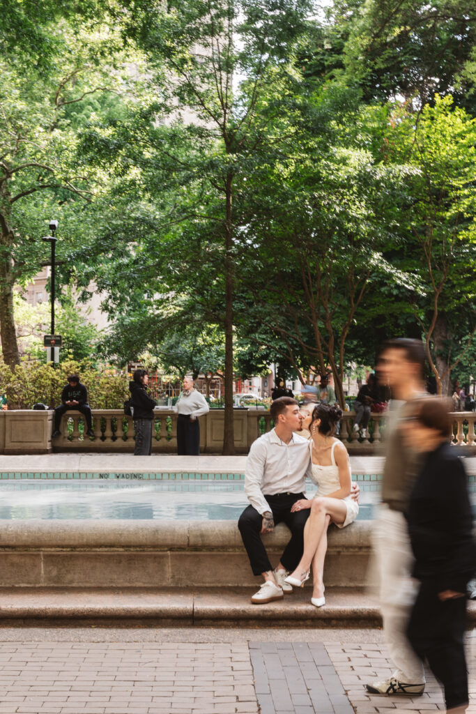 Happy couple sitting on the edge of a fountain in Rittenhouse square as people walk past in a blur, photographed by Philadelphia and New Jersey based travel wedding photographer, Madison Neumann Photography.