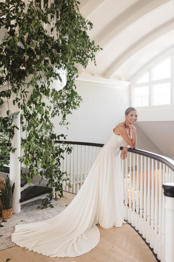 Portrait of the bride leaning on the railing in the upstairs portion of LBI National with a beautiful black and white staircase full of greenery behind her, photographed by Philadelphia and New Jersey based travel wedding photographer, Madison Neumann Photography.