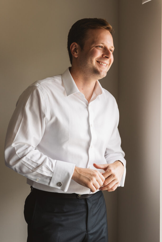 Groom in his button down smiling and looking away from the camera as he gets ready, photographed by Philadelphia and New Jersey based travel wedding photographer, Madison Neumann Photography.