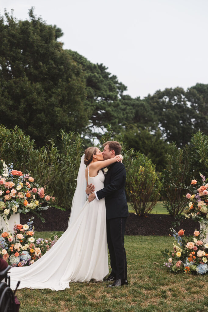 First kiss as husband and wife surrounded by gorgeous flower arrangements in shades of blue, orange, yellow, white, and pale pink, photographed by Philadelphia and New Jersey based travel wedding photographer, Madison Neumann Photography.