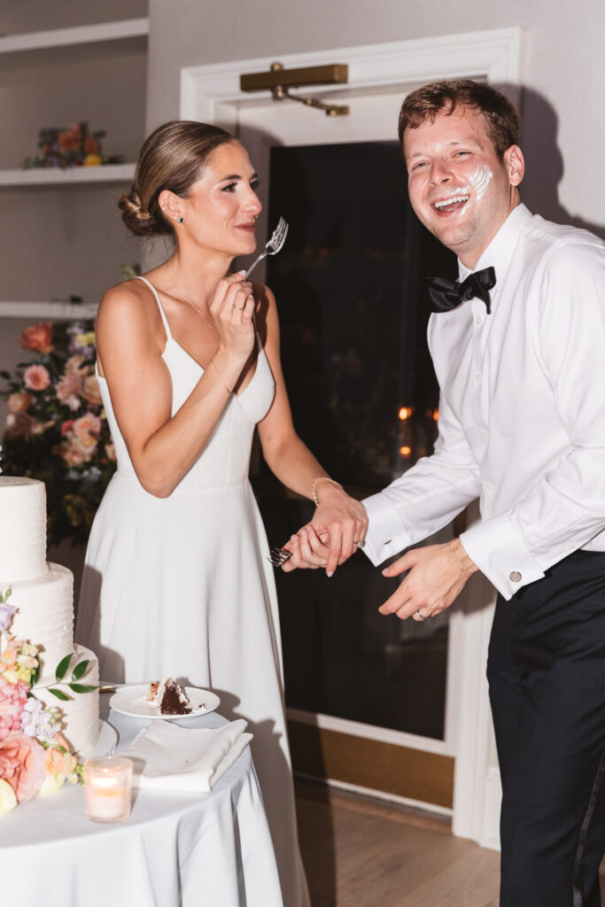 Bride happily holding the cake cutting fork after smearing cake on her husbands face as he laughs while looking at the camera, photographed by Philadelphia and New Jersey based travel wedding photographer, Madison Neumann Photography.